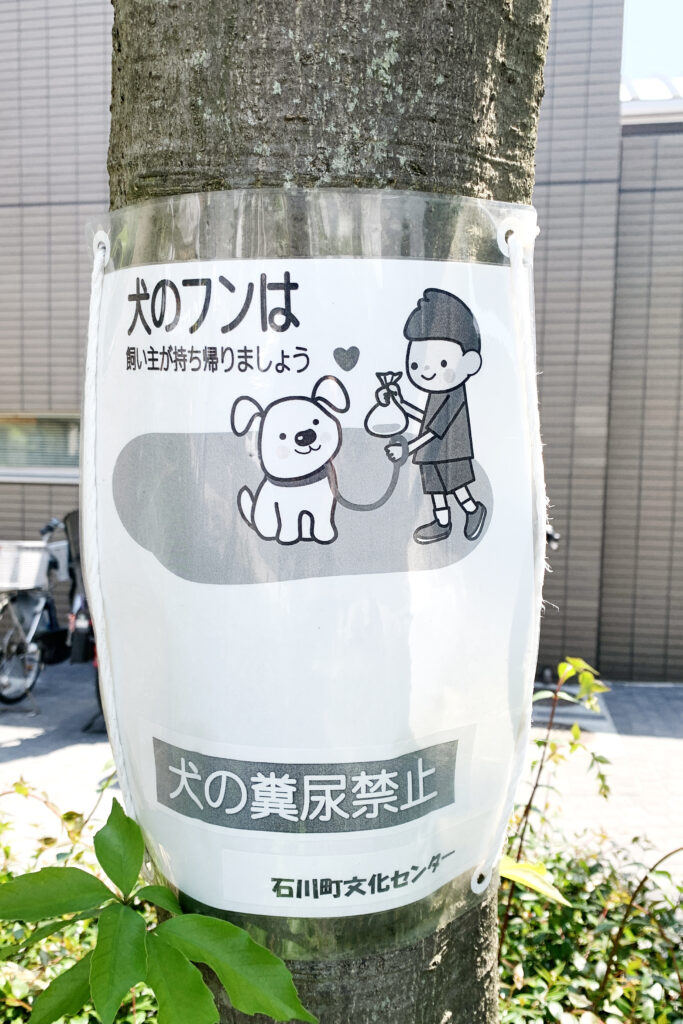 Sign on a tree near  Ishikawa Cho Culture Center reads: reads at top: Take your dog's waste home. Center: cartoon of a boy holding a bag of dog's wast, and his dog on a leash looking at him with a heart mark between them. Underneath: Dog pee prohobitied. –Ishikawa Culture Center.